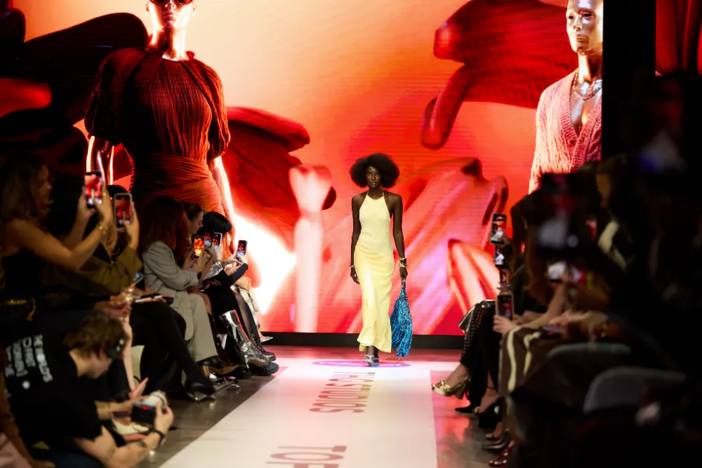 A photograph o a dark-skinned model in a yellow dress walking down a runway with a large screen showing red and orange flowing shapes.