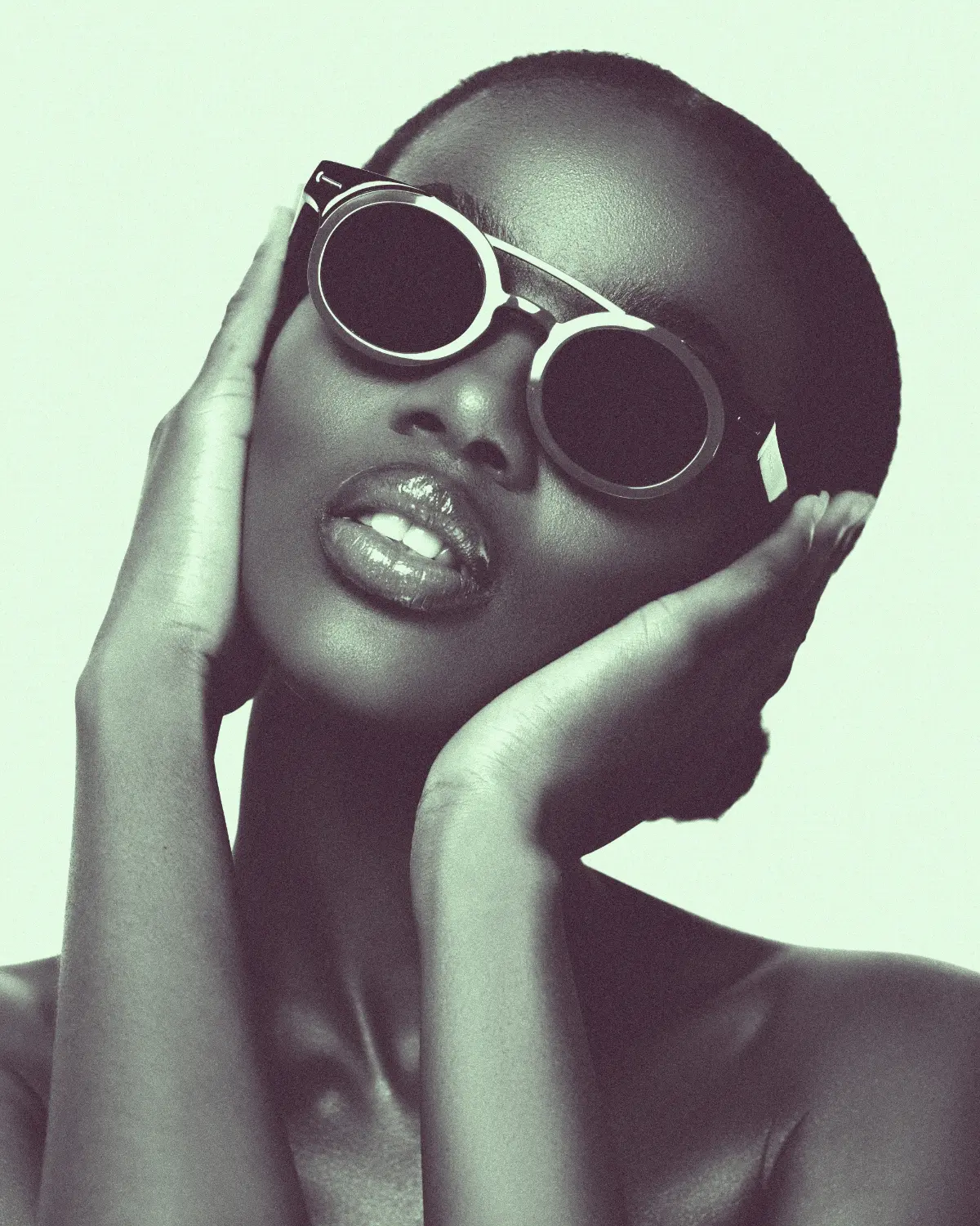 A beautiful model touches her face and leans her head to the side. She wears gold-rimmed round sunglasses.