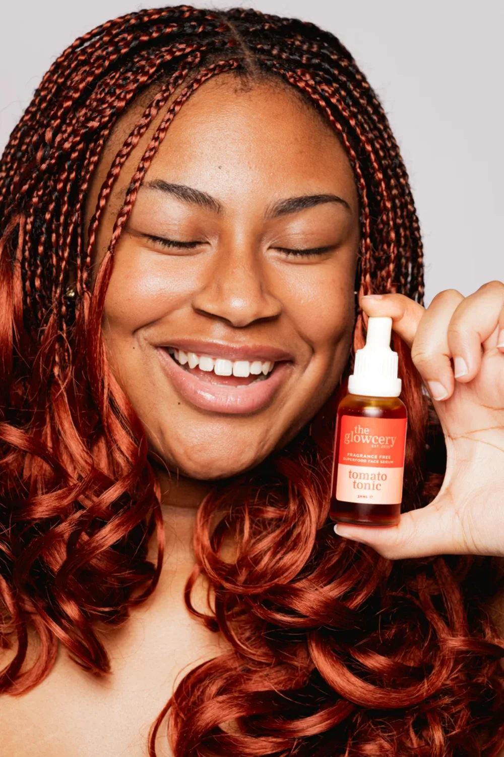 A female model smiles with her eyes closed while holding up a bottle of The Glowcery in one hand.
